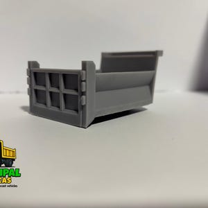 Op de afbeelding: Een grijze diecast kiepwagenbak, een miniatuurreplica van een bouwmachine. De achterkant heeft een rechthoekig ontwerp. Het logo "Municipal Replicas" staat linksonder.