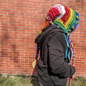 May include: A colorful, handmade crochet hat with a rainbow design. The hat features a pointed top and long, braided tassels. The hat is made of various colors, including red, pink, yellow, green, and blue.