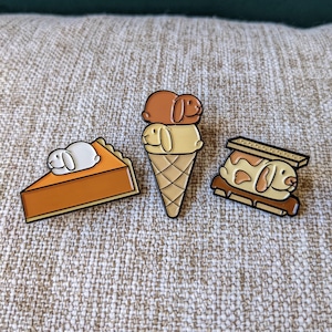 May include: Three enamel pins featuring a bunny rabbit on a pumpkin pie, ice cream cone, and s'more. The bunny is white with brown eyes and a pink nose. The background is a beige fabric.