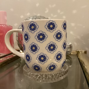 May include: White ceramic mug with a blue and yellow floral pattern. The mug is sitting on a clear glass coaster.