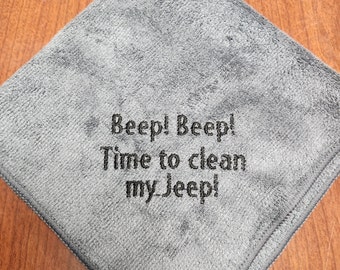 May include: A square gray microfiber cleaning cloth featuring dark gray embroidered text that reads Beep! Beep! Time to clean my Jeep! The fabric has a soft, textured pile and is finished with a stitched border along the edges of the cloth.