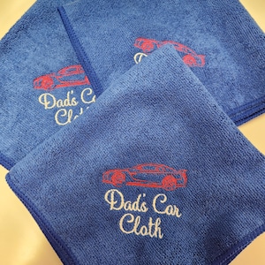 May include: Three blue microfiber cloths featuring embroidered red sports car outlines and the text "Dad's Car Cloth" in white script lettering. Each square cloth has a matching blue stitched border and a soft, textured surface.