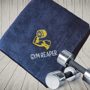 Può includere: Un asciugamano da palestra quadrato blu navy con bordo nero. L'asciugamano presenta un ricamo giallo raffigurante un braccio flesso che regge un manubrio, sopra la scritta bianca ricamata GYM REAPER, accompagnato da un manubrio in metallo.