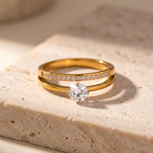 Puede incluir: Un anillo de tono dorado con un diseño de doble banda. La banda superior está incrustada con una fila de pequeñas piedras transparentes de corte redondo, mientras que la banda inferior muestra una piedra transparente redonda más grande.