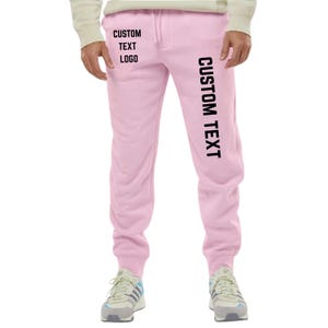 May include: Light pink sweatpants with the text "CUSTOM TEXT LOGO" printed on the left leg and "CUSTOM TEXT" printed vertically on the right leg. The sweatpants have a drawstring and are worn with sneakers.