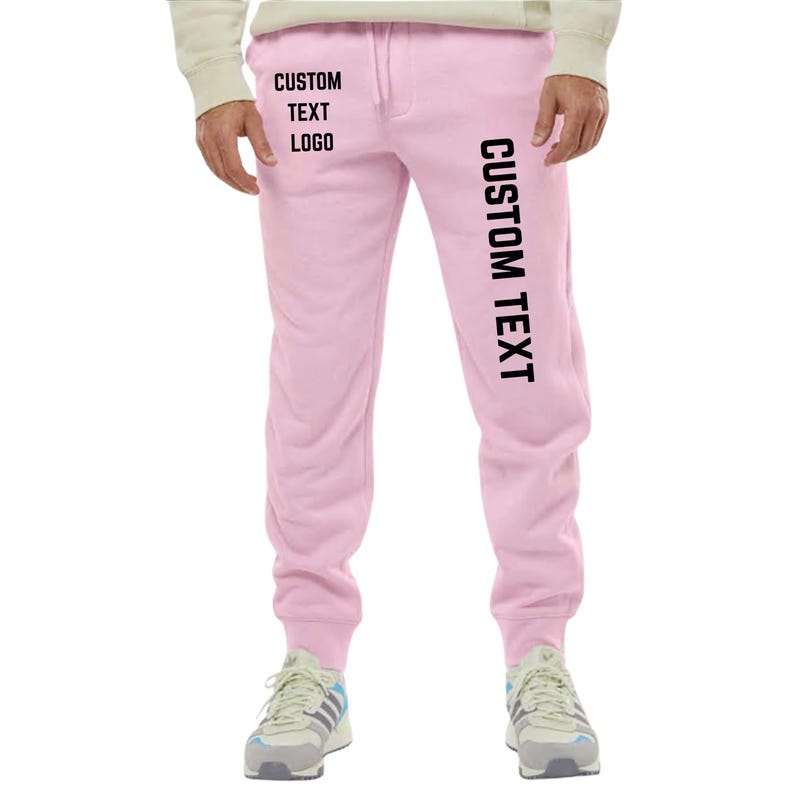 May include: Light pink sweatpants with the text "CUSTOM TEXT LOGO" printed on the left leg and "CUSTOM TEXT" printed vertically on the right leg. The sweatpants have a drawstring and are worn with sneakers.