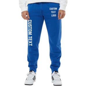 May include: Royal blue sweatpants with white text that reads "CUSTOM TEXT" vertically down the left leg and "CUSTOM TEXT LOGO" on the right leg. The sweatpants have a drawstring waist and cuffed ankles. The person is wearing white sneakers.