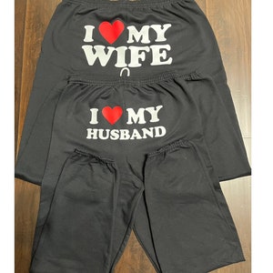May include: Two pairs of black sweatpants with white text and a red heart. The top pair says "I love my wife" and the bottom pair says "I love my husband".