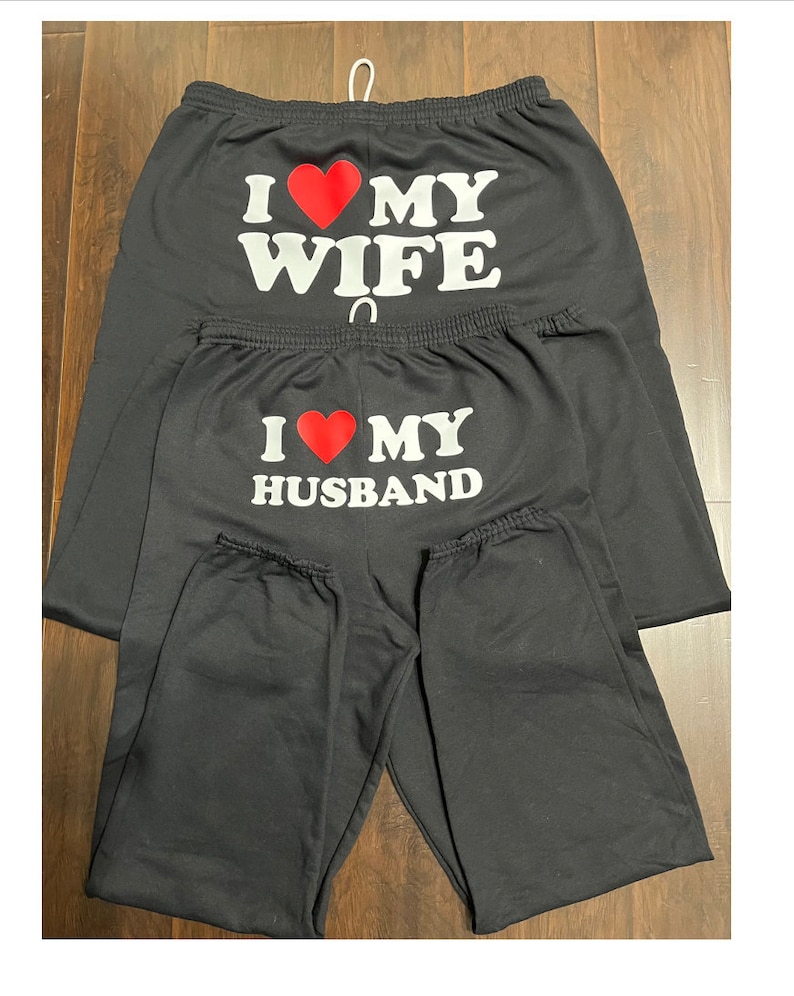 May include: Two pairs of black sweatpants with white text and a red heart. The top pair says "I love my wife" and the bottom pair says "I love my husband".