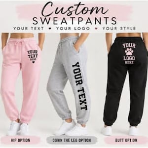 May include: Three pairs of custom sweatpants in light pink, heather gray, and black. The pink pair features text on the hip, the gray pair has vertical text down the leg, and the black pair displays a logo on the back. Each pair has an elastic waistband.