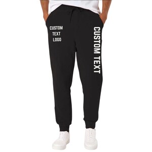 May include: Black sweatpants with a drawstring waist and white text that reads "CUSTOM TEXT LOGO" on the left leg and "CUSTOM TEXT" vertically down the right leg. The pants are shown with white sneakers.