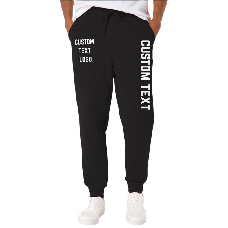 May include: Black sweatpants with a drawstring waist and white text that reads "CUSTOM TEXT LOGO" on the left leg and "CUSTOM TEXT" vertically down the right leg. The pants are shown with white sneakers.