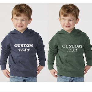 May include: Two long-sleeve hooded sweatshirts, one in heather navy blue and one in heather forest green. Both feature a front kangaroo pocket and the white text CUSTOM TEXT printed in a bold, sans-serif font across the center of the chest.