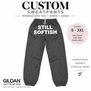 May include: Dark gray sweatpants featuring a white drawstring waist and the text STILL SOFTISH printed in white on the upper back. The garment is labeled as a Gildan Heavy Blend 18200, composed of 50% cotton and 50% polyester, available in sizes S-3XL.