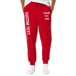 May include: Red sweatpants with white text that reads "CUSTOM TEXT" vertically down the left leg and "CUSTOM TEXT LOGO" on the right leg. The sweatpants have a drawstring and are worn with white sneakers.