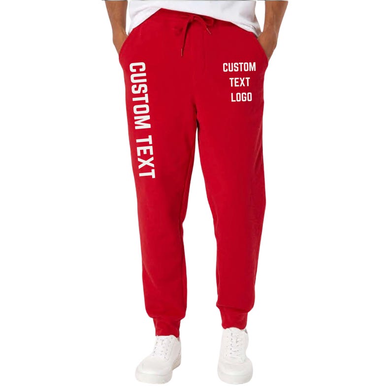May include: Red sweatpants with white text that reads "CUSTOM TEXT" vertically down the left leg and "CUSTOM TEXT LOGO" on the right leg. The sweatpants have a drawstring and are worn with white sneakers.