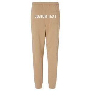 May include: Tan sweatpants with a drawstring waist and elastic cuffs. The back of the pants features a pocket and the text "CUSTOM TEXT" in white. These comfortable pants are suitable for casual wear.