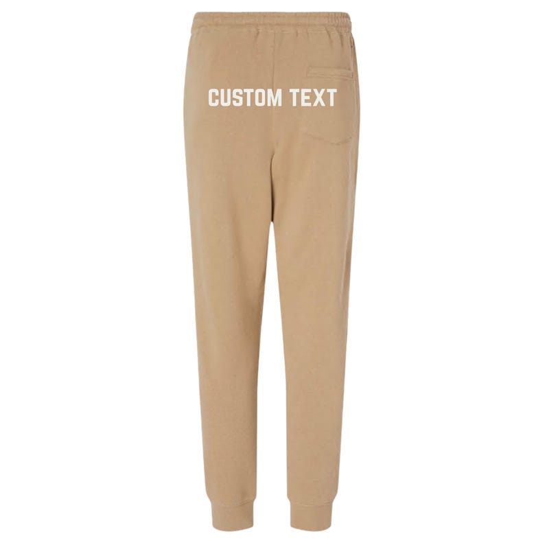 May include: Tan sweatpants with a drawstring waist and elastic cuffs. The back of the pants features a pocket and the text "CUSTOM TEXT" in white. These comfortable pants are suitable for casual wear.