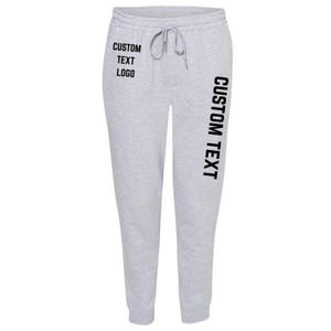 May include: Light gray sweatpants with a drawstring waist. The text "CUSTOM TEXT LOGO" is printed in black on the left leg, and "CUSTOM TEXT" is printed vertically on the right leg. The sweatpants are made of a soft material.