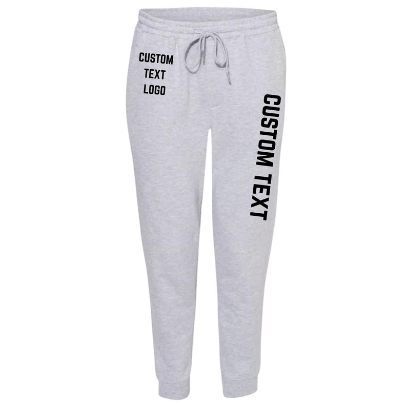 May include: Light gray sweatpants with a drawstring waist. The text "CUSTOM TEXT LOGO" is printed in black on the left leg, and "CUSTOM TEXT" is printed vertically on the right leg. The sweatpants are made of a soft material.