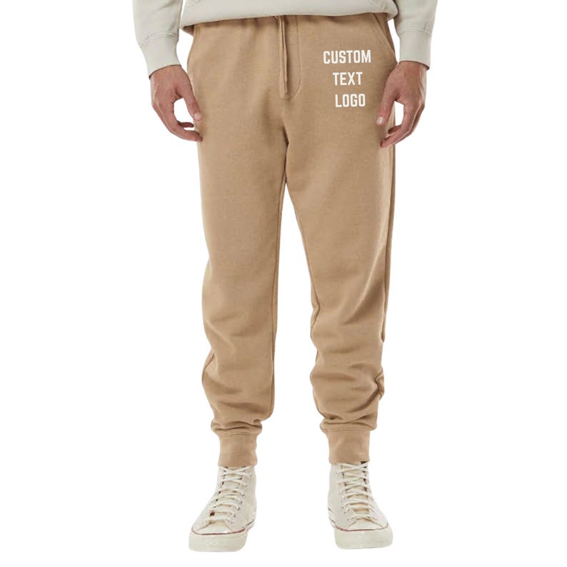 May include: Tan sweatpants with a drawstring waist and elastic cuffs. The text "CUSTOM TEXT LOGO" is printed in white on the left thigh. The person is wearing white high-top sneakers.