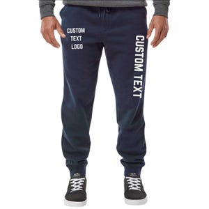 May include: Navy blue sweatpants with white text that reads "CUSTOM TEXT LOGO" on the left leg and "CUSTOM TEXT" on the right leg. The pants have a drawstring waist and are worn with black and white sneakers.