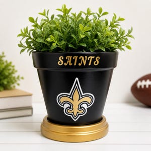 May include: Black flower pot with the word "SAINTS" in gold lettering and a gold fleur-de-lis logo. The pot is filled with green leafy plants and sits on a gold base. A football is visible in the background.