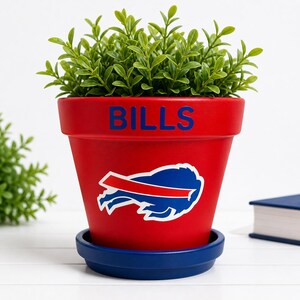 May include: A red flower pot with the word "BILLS" in blue lettering and a team logo. The pot sits on a blue saucer and contains green leafy plants. A decorative item for sports fans.