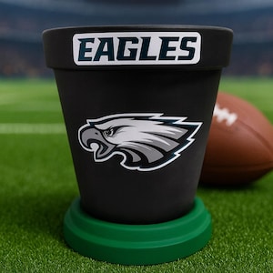May include: Black flower pot with the word "EAGLES" and a team logo. The pot sits on a green base, with a football in the background. The image is set on a green field.