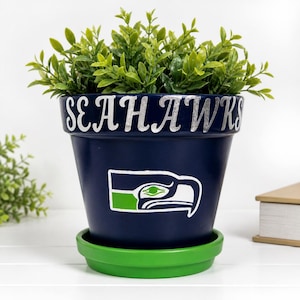 May include: A navy blue flower pot with the word "SEAHAWKS" in silver lettering. The pot features the team logo and sits on a green saucer. The pot is filled with green leafy plants.
