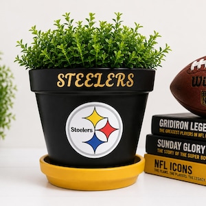 May include: A black ceramic plant pot with a yellow saucer, featuring the word Steelers in gold lettering and the Pittsburgh Steelers logo on the front. The pot contains a green plant and is positioned next to a brown football and three books.
