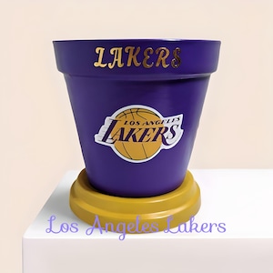 May include: A purple flower pot with the word "LAKERS" in gold lettering. The Los Angeles Lakers logo is on the front. The pot sits on a yellow base. The words "Los Angeles Lakers" are at the bottom.