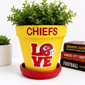 May include: A bright yellow flower pot with a red saucer, featuring the word CHIEFS in red at the top and the word LOVE in red with a Kansas City Chiefs football helmet replacing the letter O. A green plant sits inside the pot.