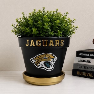 May include: A black ceramic flower pot featuring the gold text JAGUARS and a jaguar head logo. The pot sits on a gold saucer and contains a green artificial plant. Beside it are stacked books labeled Jacksonville Jaguars, Jaguars, 100 Seasons, and DUUUVAL.
