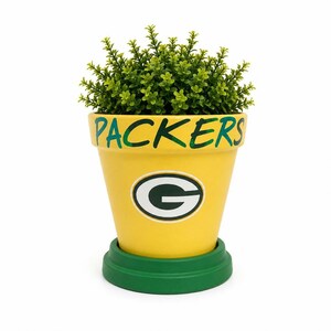 May include: A yellow terracotta plant pot with a green base and rim. The word PACKERS is painted in green on the rim, and a white and green G logo is centered on the front. A small, bushy green artificial plant sits inside the pot.