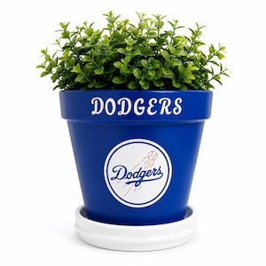 May include: A blue flower pot with a white saucer containing a green plant. The pot features the white text DODGERS at the top and a circular white logo with the word Dodgers and a baseball graphic in the center on the front of the blue surface.