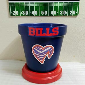 May include: A blue Bills-themed flower pot with a red base. The pot features the word "BILLS" in red and a heart-shaped logo. A welcome sign with a football field design is in the background.