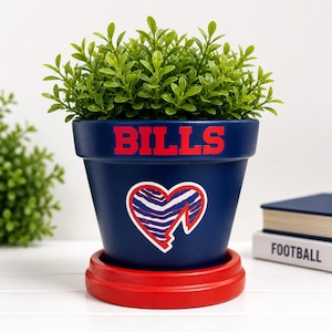 May include: A navy blue flower pot with the word "BILLS" in red. The pot features a heart-shaped logo with a red outline and a blue and white zebra stripe design. The pot sits on a red saucer and contains a green plant.