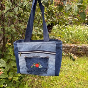 Könnte beinhalten: Eine Tragetasche aus hell- und dunkelblauem Jeansstoff mit zwei Schulterriemen, einer horizontalen Reißverschlusstasche und einer aufgesetzten Außentasche, die mit einer kleinen, bunten Blumenstickerei in Rot, Grün und Gelb verziert ist.