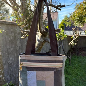 May include: A rectangular tote bag featuring a patchwork design of various fabric panels in shades of brown, tan, gray, and olive green. The bag includes a horizontal zippered pocket on the front with a gold-toned zipper pull and two brown straps.