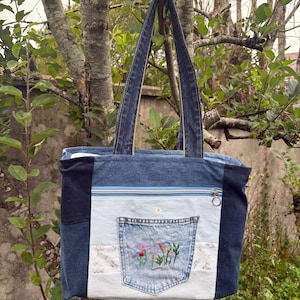 Può includere: Una borsa tote realizzata con varie tonalità di tessuto denim blu. La borsa presenta una tasca orizzontale con cerniera, due tracolle e una tasca centrale applicata decorata con ricami floreali colorati in rosso, giallo e verde.