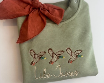 May include: A green sweatshirt with a brown bow on top. The sweatshirt has three embroidered ducks and the name "Lila James" embroidered below the ducks.