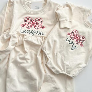 Op de afbeelding: Twee cr&egrave;mekleurige babyoutfits. E&eacute;n is een jurk met lange mouwen en zakken, geborduurd met een rode strik en de naam "teagan". De andere is een onesie met een vergelijkbare strik en de naam "ivy".