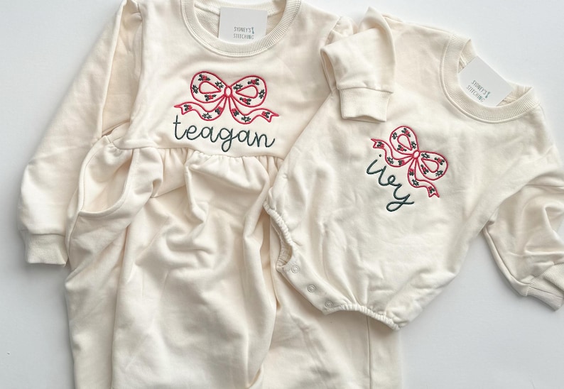 Op de afbeelding: Twee cr&egrave;mekleurige babyoutfits. E&eacute;n is een jurk met lange mouwen en zakken, geborduurd met een rode strik en de naam "teagan". De andere is een onesie met een vergelijkbare strik en de naam "ivy".