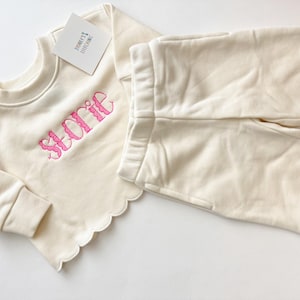 May include: Cream-colored baby outfit set featuring a crew neck sweatshirt and matching pants. The sweatshirt has the name "Storie" embroidered in pink. Both pieces have a scalloped edge. A tag reads "Storie's Stitching".
