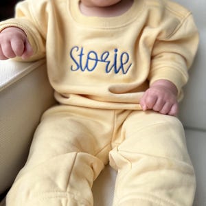 Può includere: Un bambino indossa una tuta gialla chiara composta da felpa e pantaloni abbinati. La felpa ha il nome "Storie" ricamato in blu sul petto. L'outfit sembra essere realizzato in un materiale morbido e confortevole.