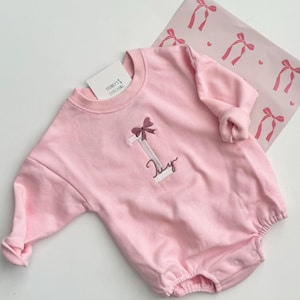 May include: Pink baby onesie with a white letter I and a pink bow embroidered on the front. The onesie has a white text that says "Lily".
