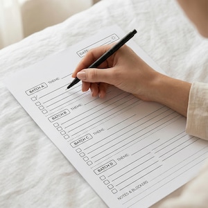 May include: A white paper planner with a black pen. The planner has sections labeled "BATCH A," "BATCH B," "BATCH C," and "BATCH D," with space for themes and notes. The word "DATE" is at the top. The pen is held by a person.