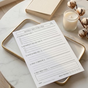 May include: A white Weekly Batch Review notepad with a search bar, checkboxes, and prompts for notes. The notepad is on a gold-rimmed tray, with a book, candle, and decorative cotton stems in the background. The notepad includes a mood tracker.