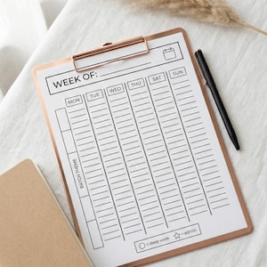 May include: A rose gold clipboard with a weekly planner sheet, a black pen, and a small tan notebook. The planner sheet has sections for each day of the week, with the text "WEEK OF:" at the top. The clipboard is on a white surface.
